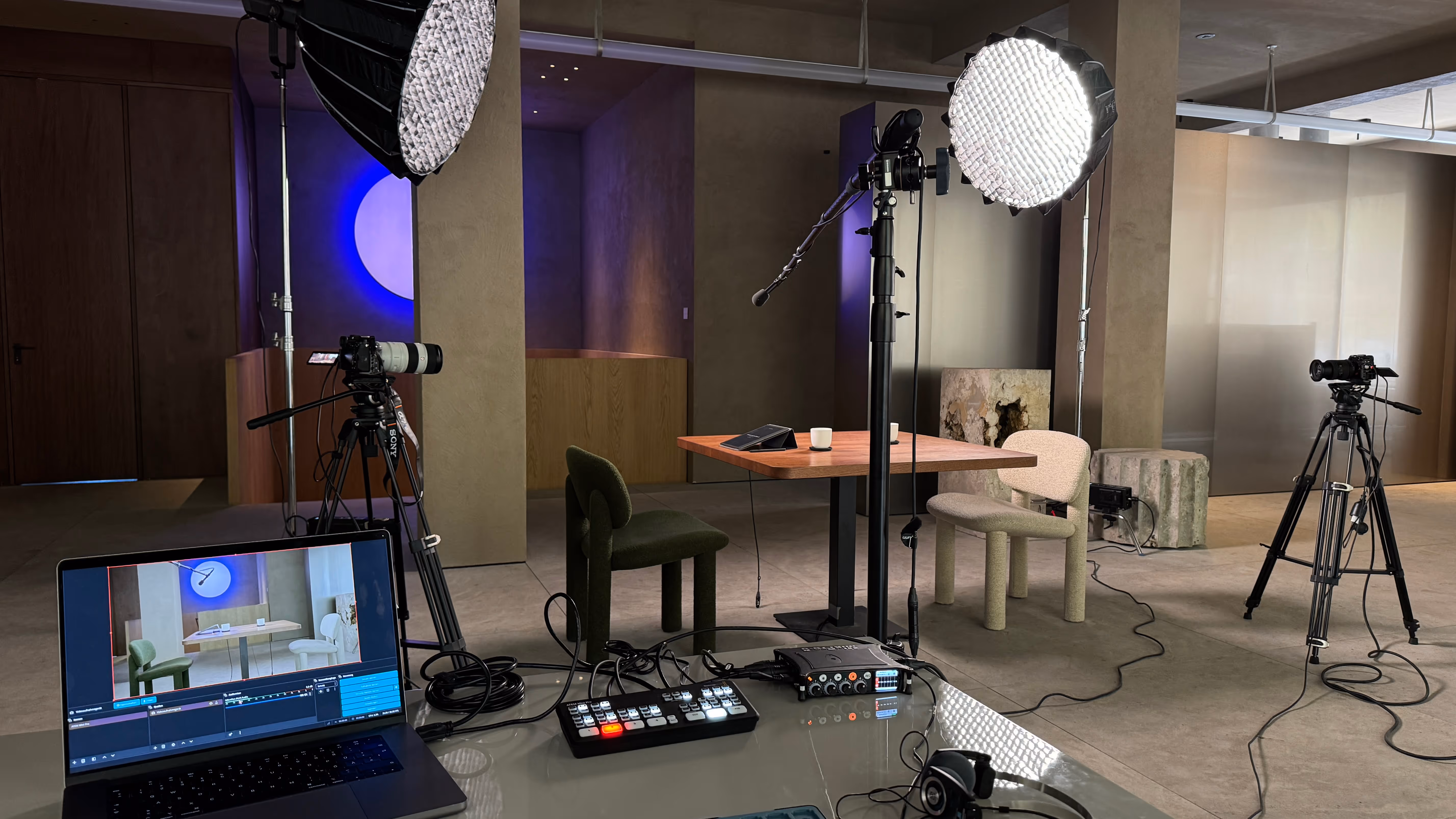 Professional video setup with two cameras on tripods, studio lights, a table with two chairs, and a laptop showing live video feed in a modern room.