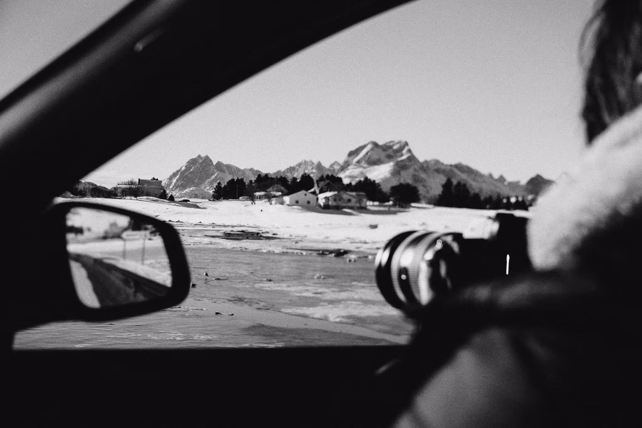 Person inside a car holding a camera pointing at a snowy landscape with houses and mountains in the background.