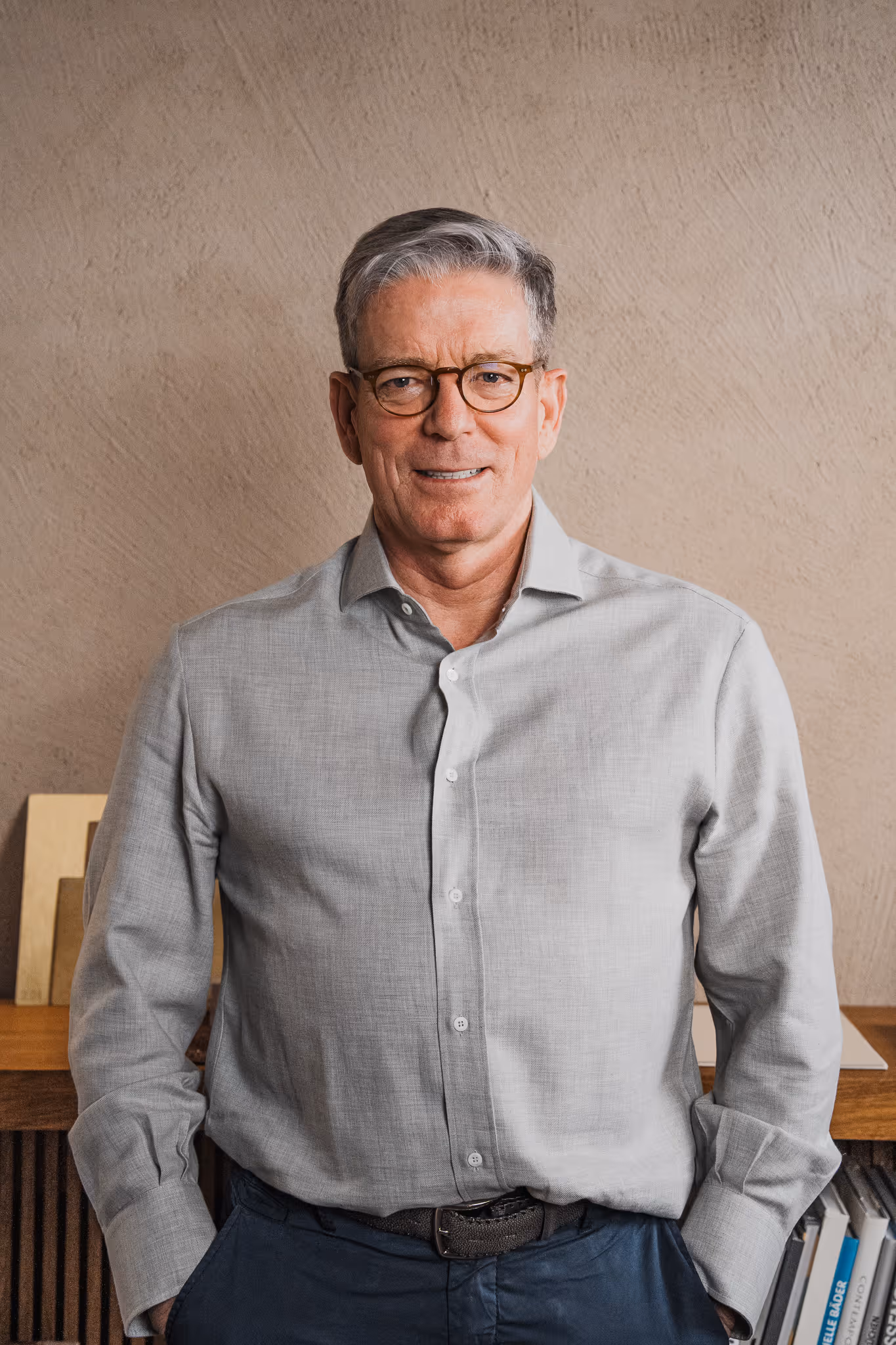 Smiling middle-aged man with gray hair and glasses wearing a light gray button-up shirt and dark pants, standing indoors.