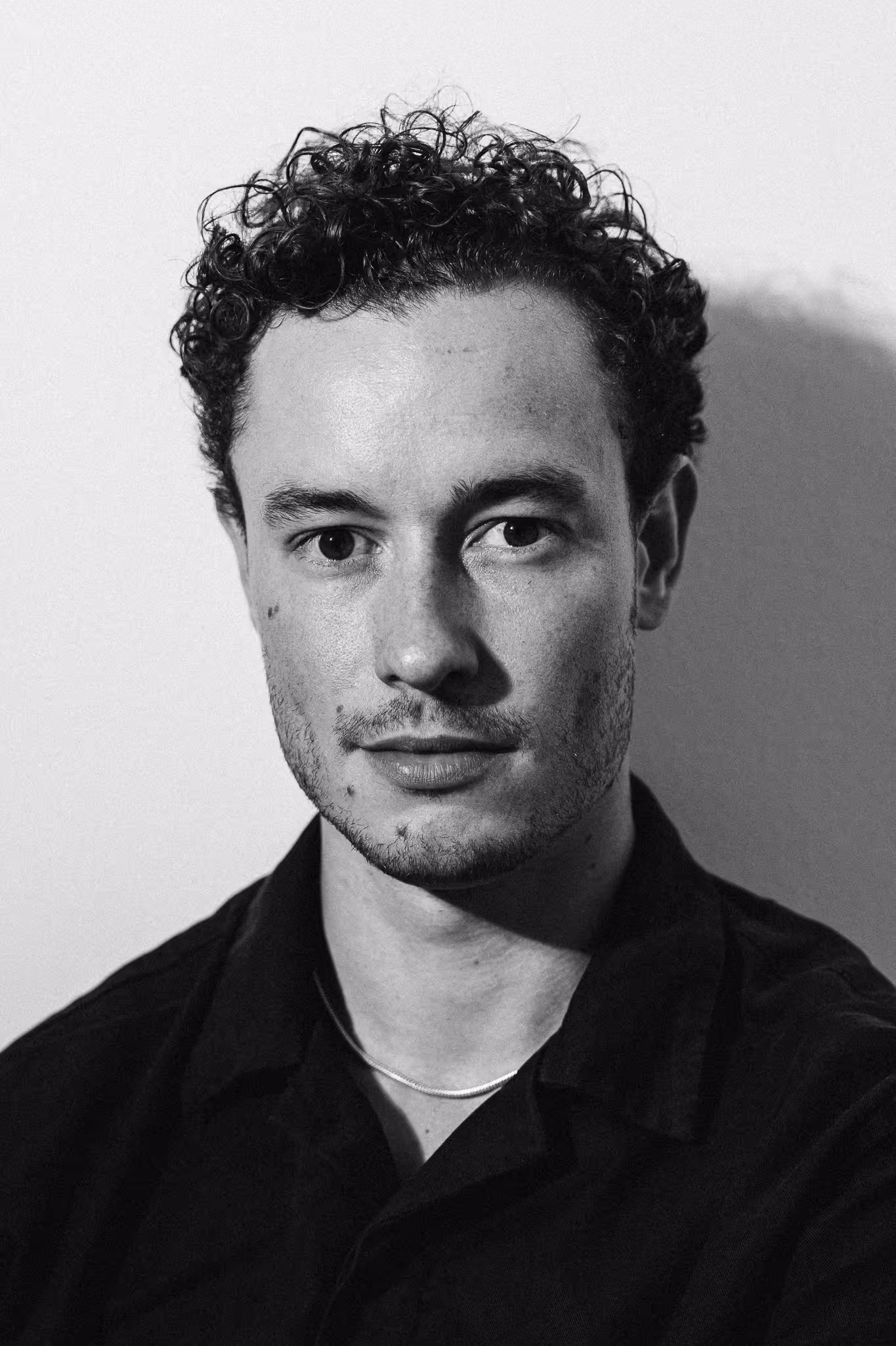 Black and white close-up portrait of a man with curly hair and light facial stubble wearing a dark shirt and a thin necklace.