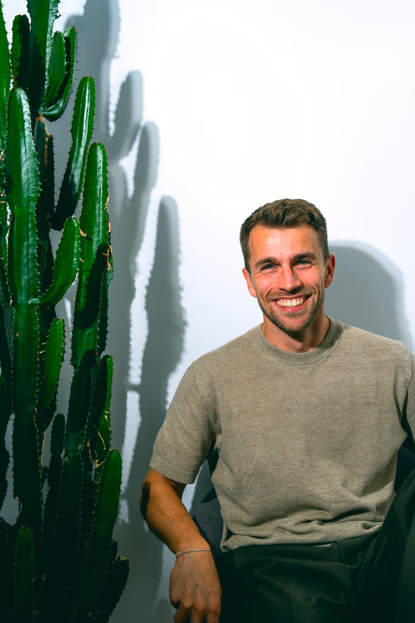 Smiling man sitting beside a tall green cactus casting shadows on a white wall.