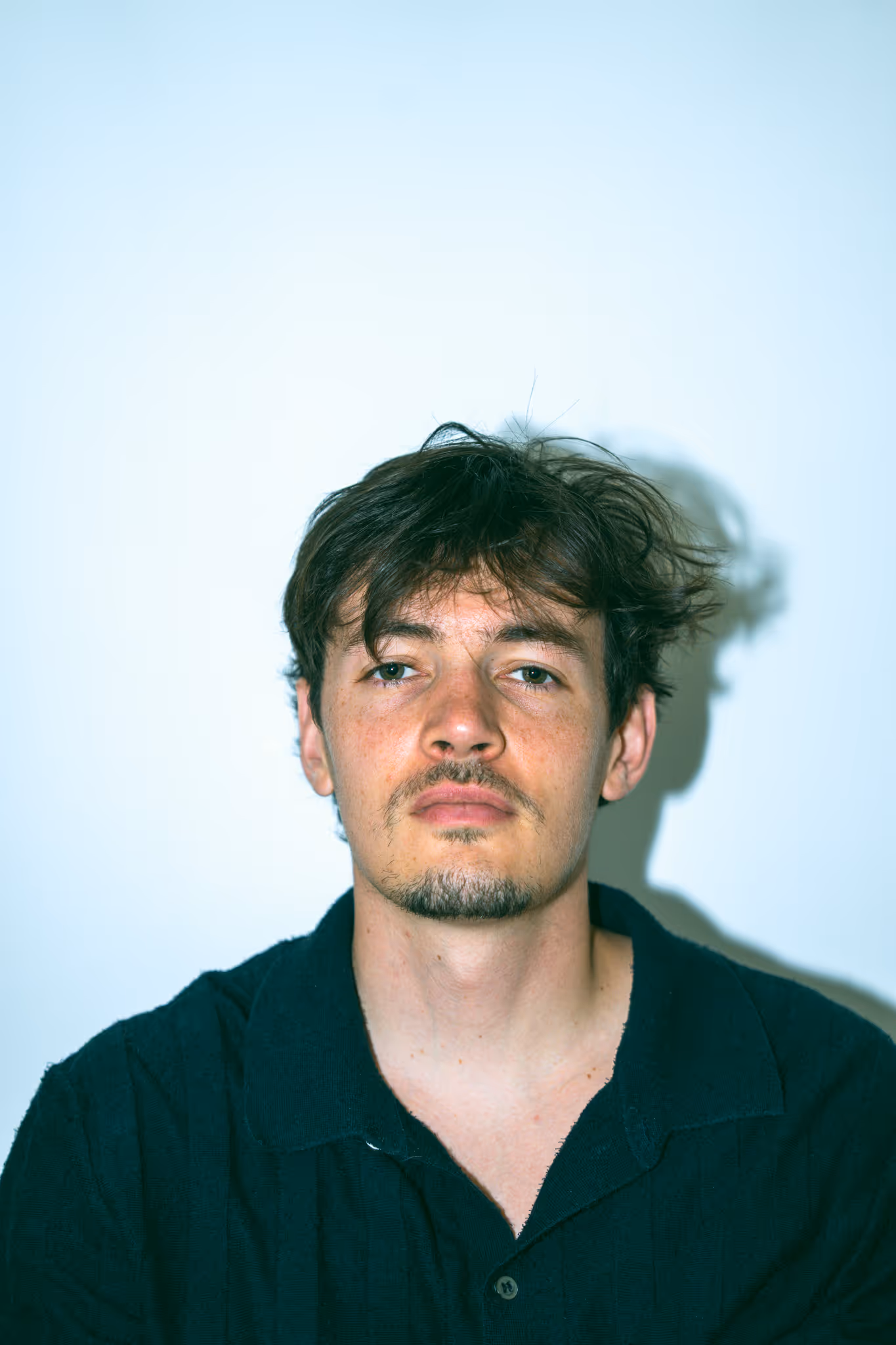 Portrait of a young man with messy dark hair and facial hair wearing a black collared shirt against a plain light background.