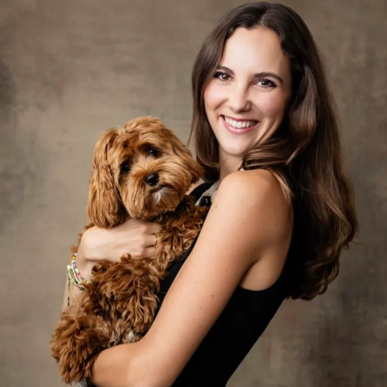 Smiling woman with long brown hair holding a small fluffy brown dog against a neutral background.