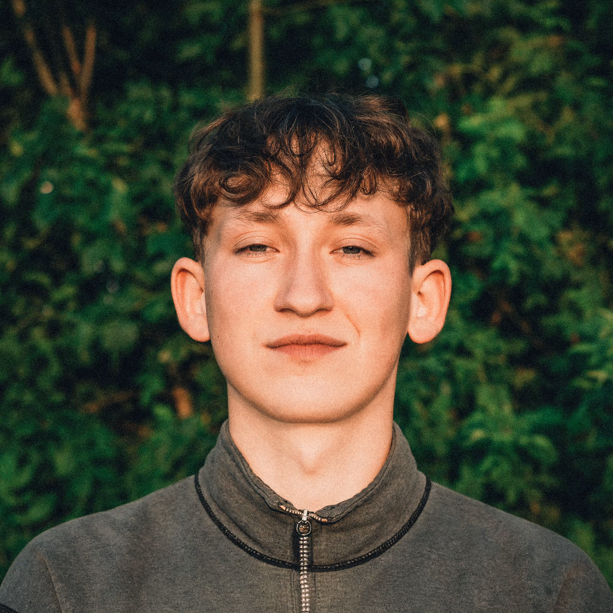 Young man with short curly brown hair wearing a gray zip-up jacket, standing in front of green leafy background.