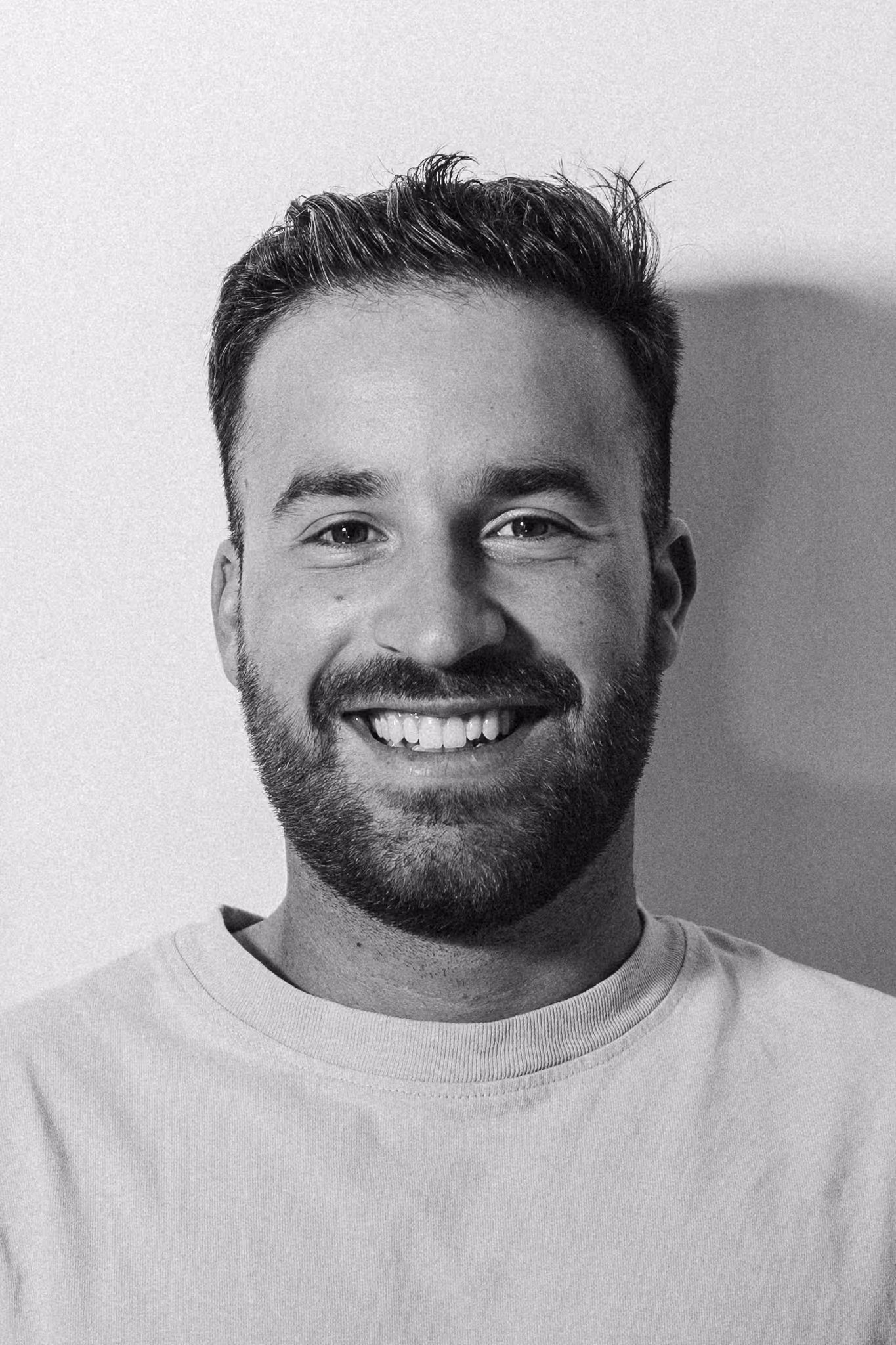 Smiling bearded man with short hair wearing a light-colored crew neck shirt against a plain background.