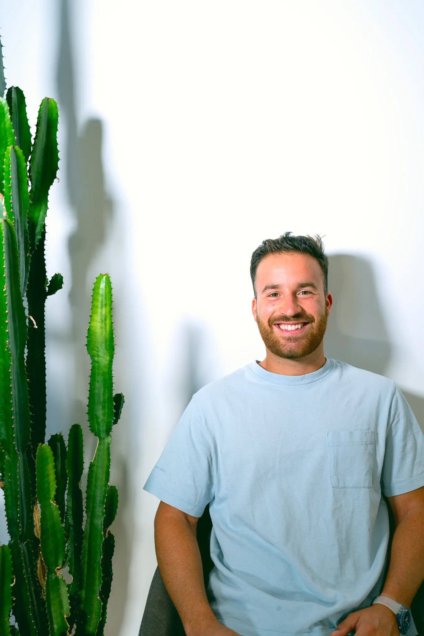 Smiling man with beard in a light blue t-shirt sitting near tall green cactus against white wall.