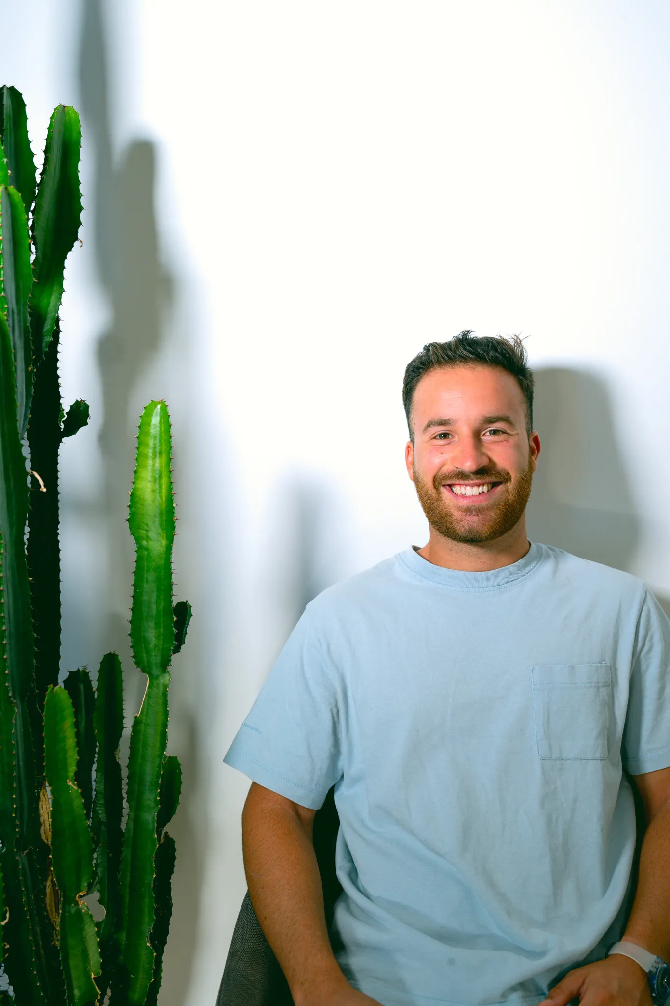 Smiling man with beard wearing a light blue t-shirt sitting next to a tall green cactus against a white wall.