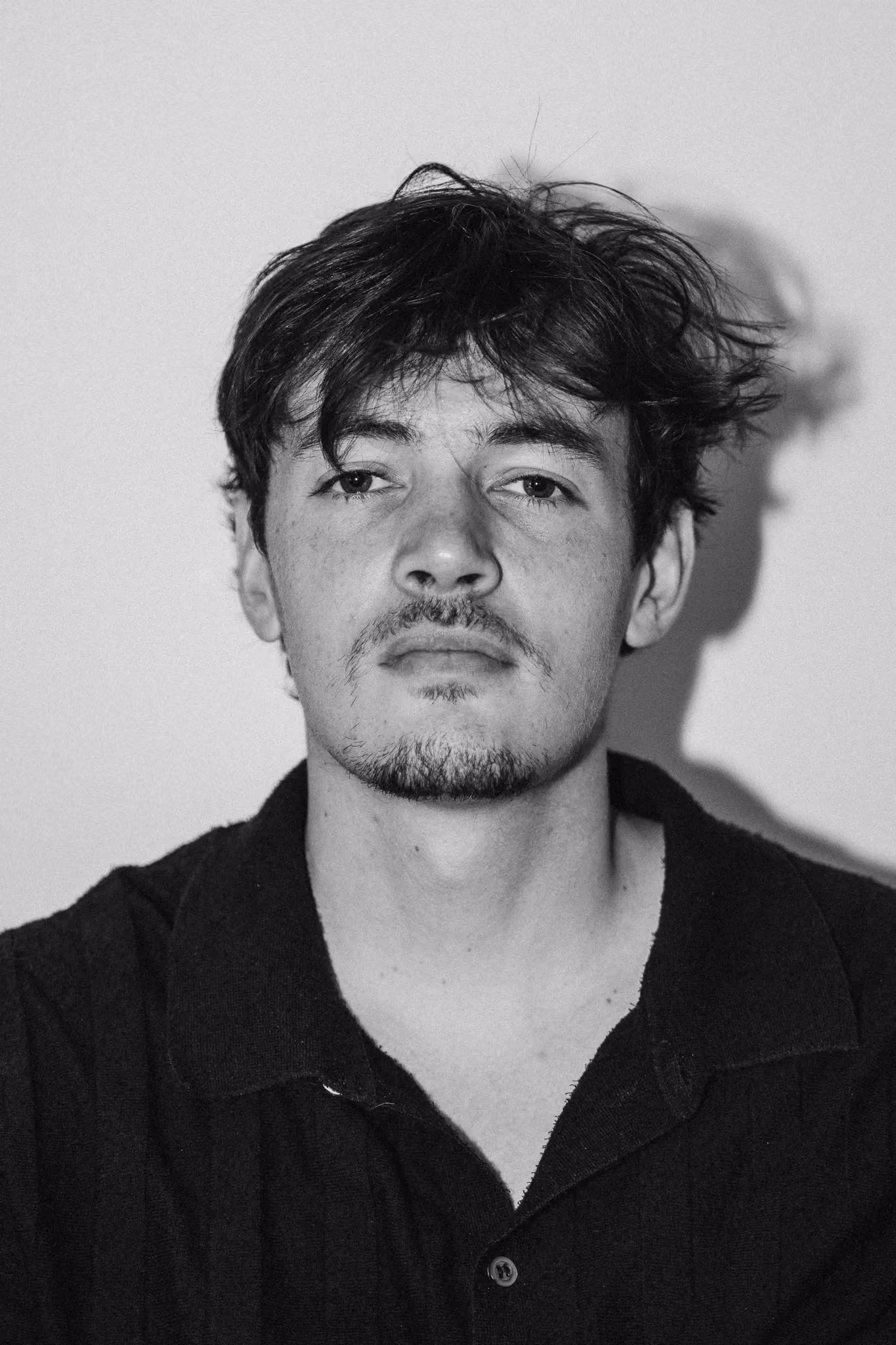 Black and white portrait of a young man with tousled hair and light facial hair wearing a dark collared shirt against a plain background.