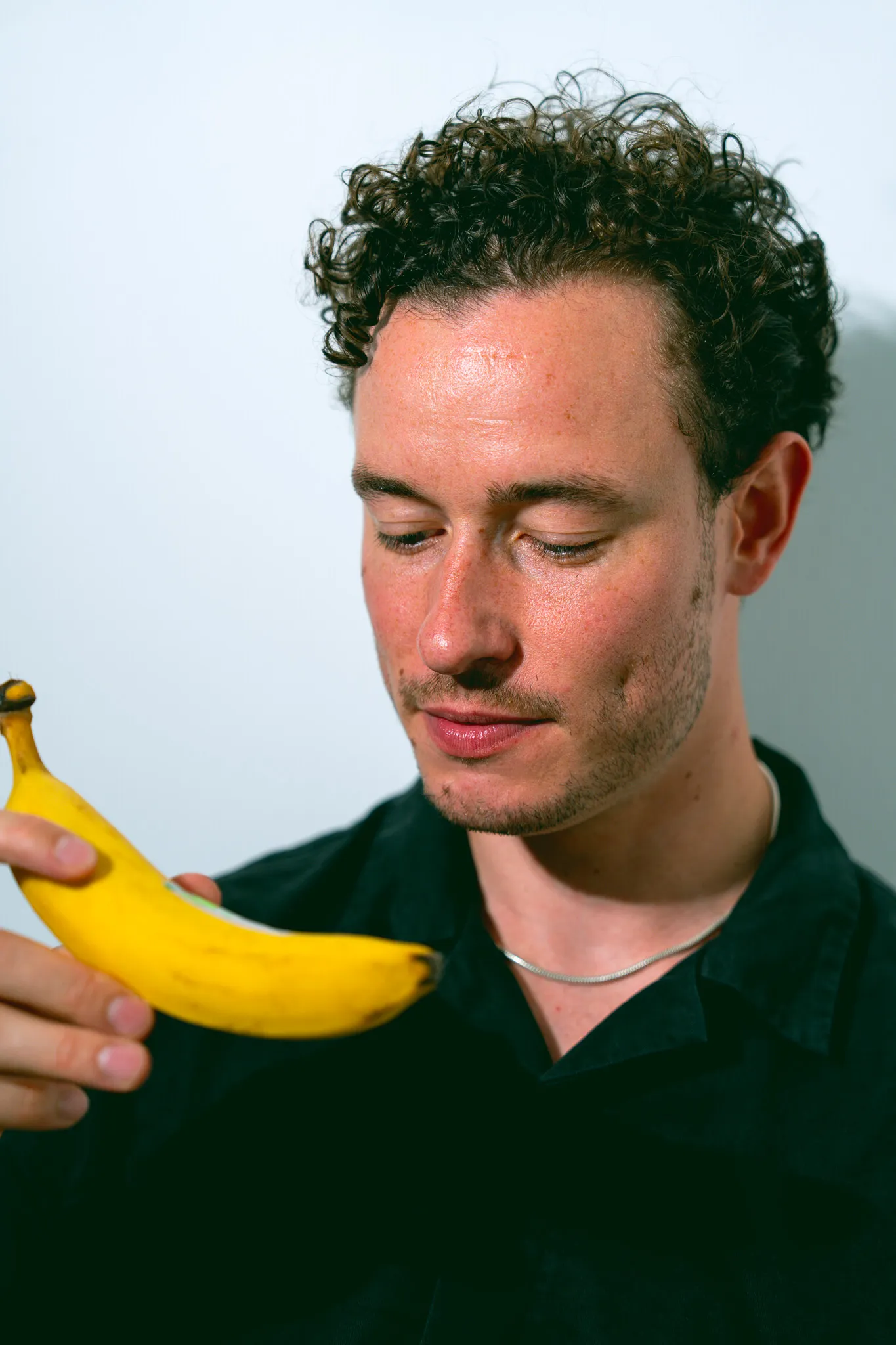 Young man with curly hair looking thoughtfully at a banana he is holding.