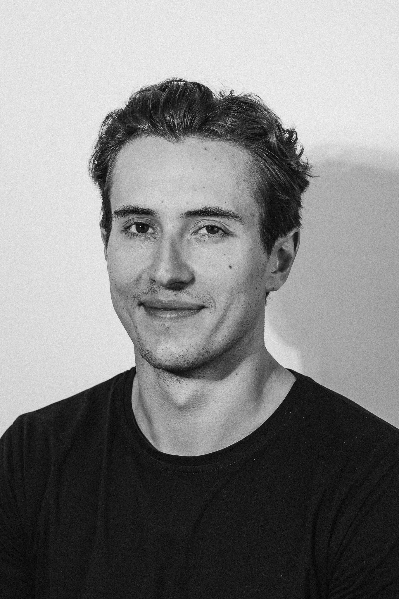 Black and white portrait of a young man with short hair wearing a black shirt, smiling slightly against a plain background.