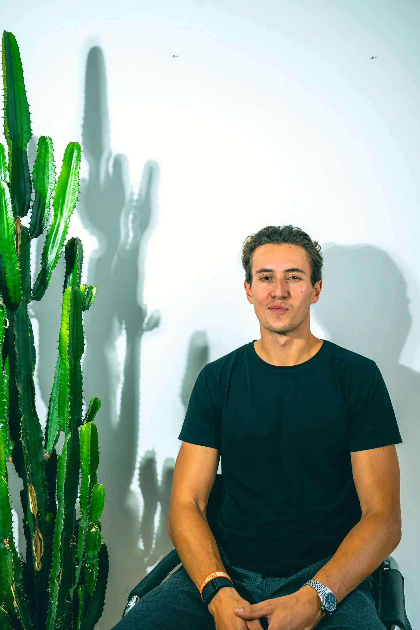 Young man in a black t-shirt sitting next to a tall green cactus casting a shadow on a white wall.