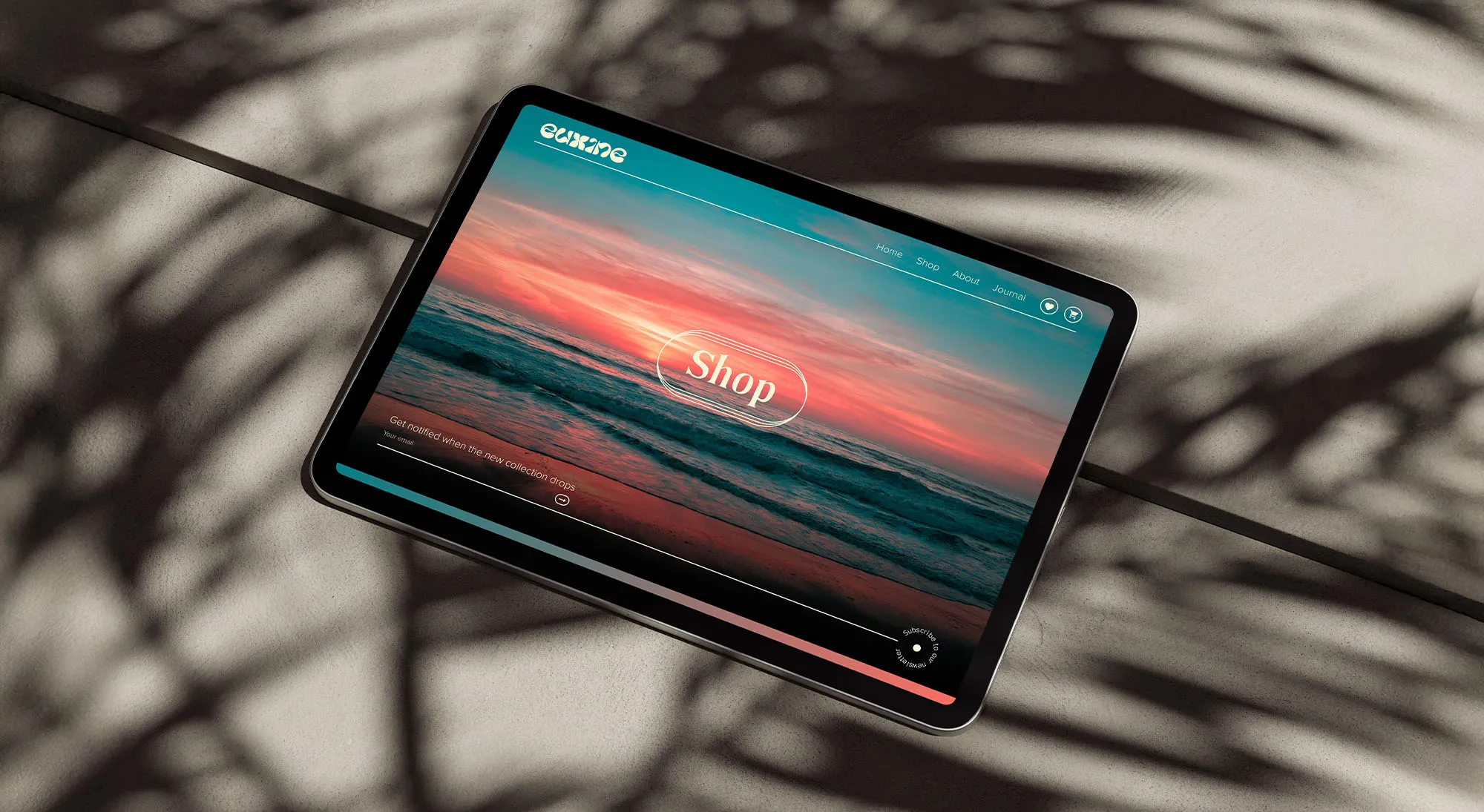 Tablet displaying a website homepage with a sunset ocean background and a central 'Shop' button, surrounded by shadows of palm leaves.