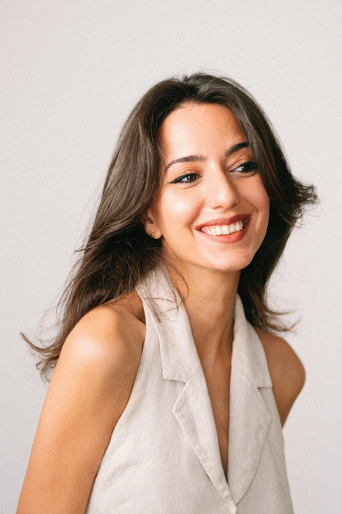 Smiling young woman with long brown hair, wearing a sleeveless beige top, looking to the side against a plain background.