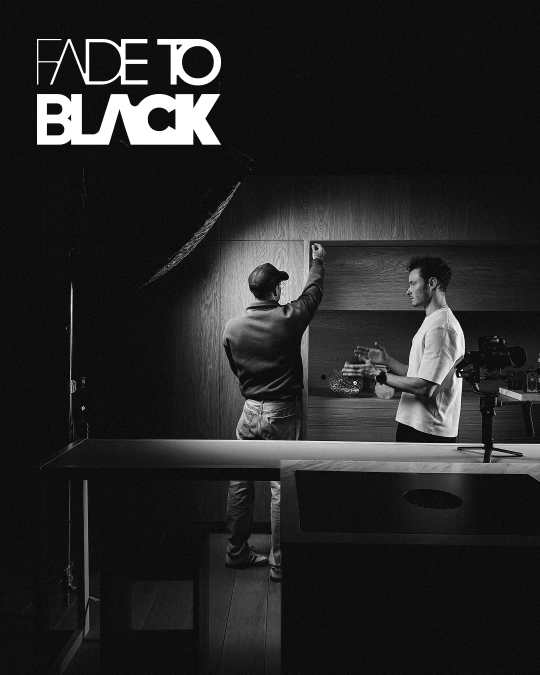 Two men working in a dimly lit room with one adjusting a shelf and the other gesturing near a camera setup.