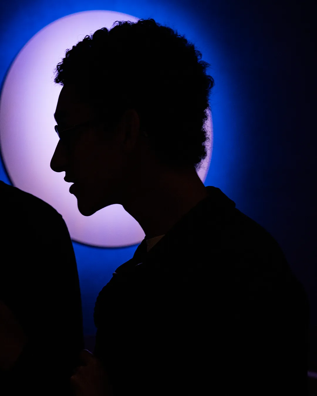 Silhouette of a person with curly hair and glasses against a blue background with a large circular light.