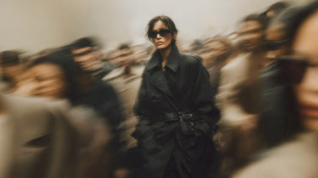 Woman in a black trench coat and sunglasses walking through a crowd with blurred motion effect.