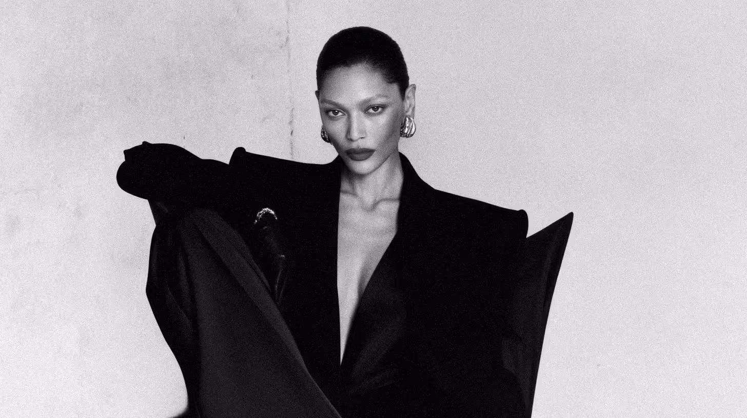 Black and white photo of a woman with slicked-back hair wearing statement earrings and a dramatic oversized blazer with sharp shoulders, looking directly at the camera.