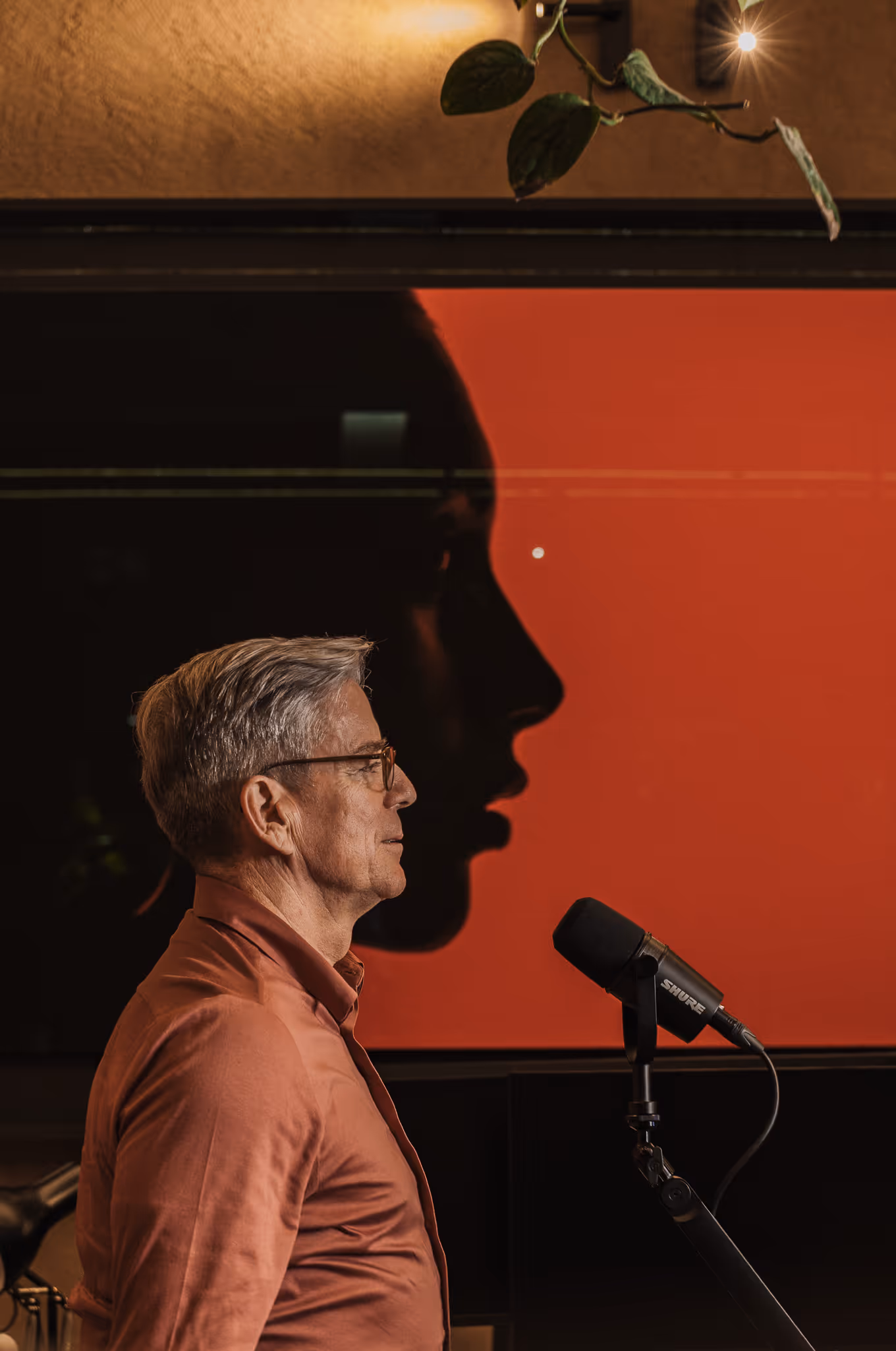 man speaking into a podcast microphone against a background featuring a large red screen 