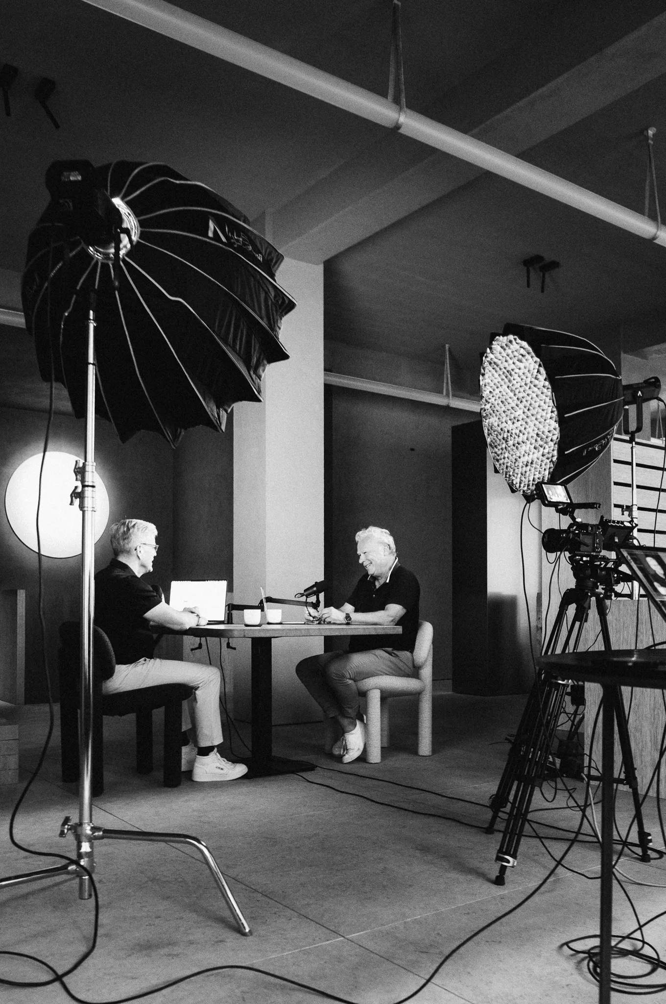 Two men sitting at a table in a studio setting with professional lighting and camera equipment around them.