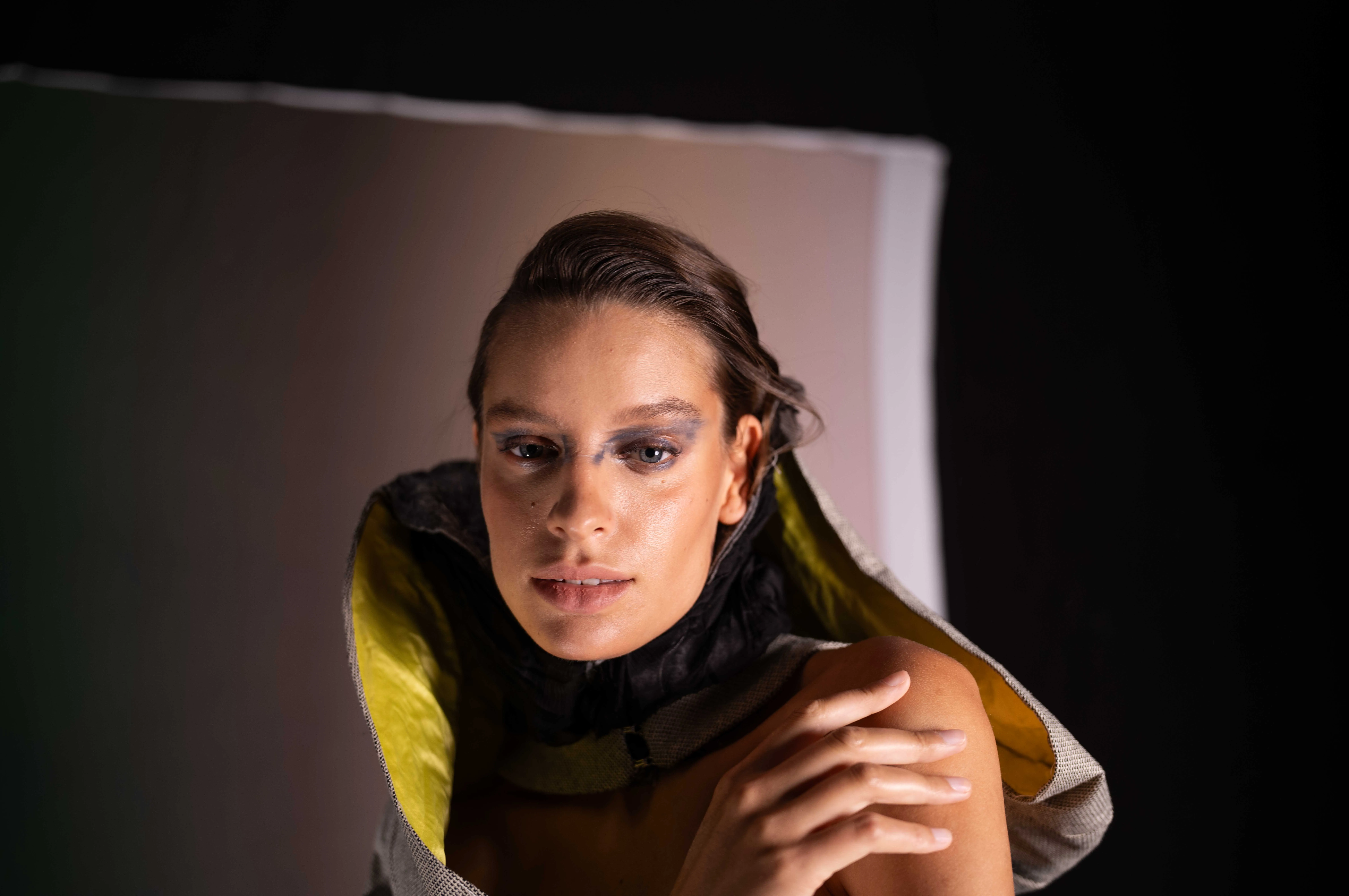 Portrait of a woman with wet-look hair, dark smoky eye makeup, and a high-collared garment with yellow lining, touching her shoulder.