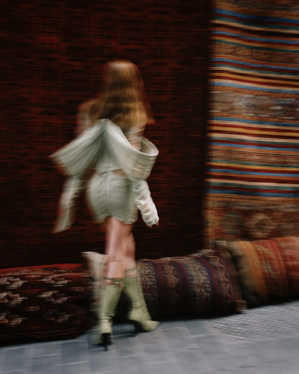 Blurred image of a woman in a green outfit and boots walking past colorful patterned rugs and cushions.