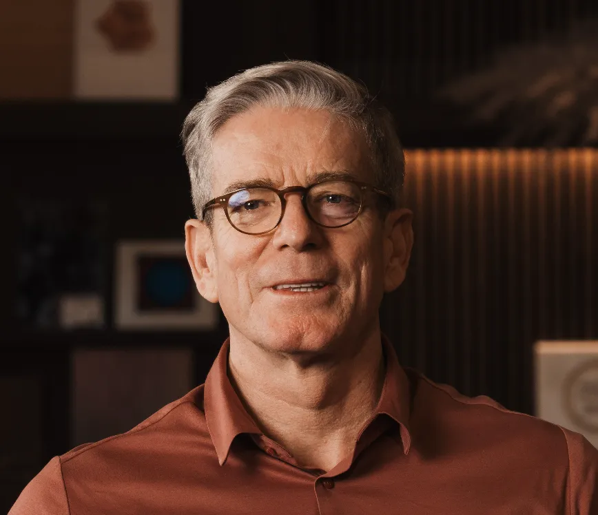 Middle-aged man with gray hair and glasses wearing a rust-colored shirt standing indoors with a blurred background.