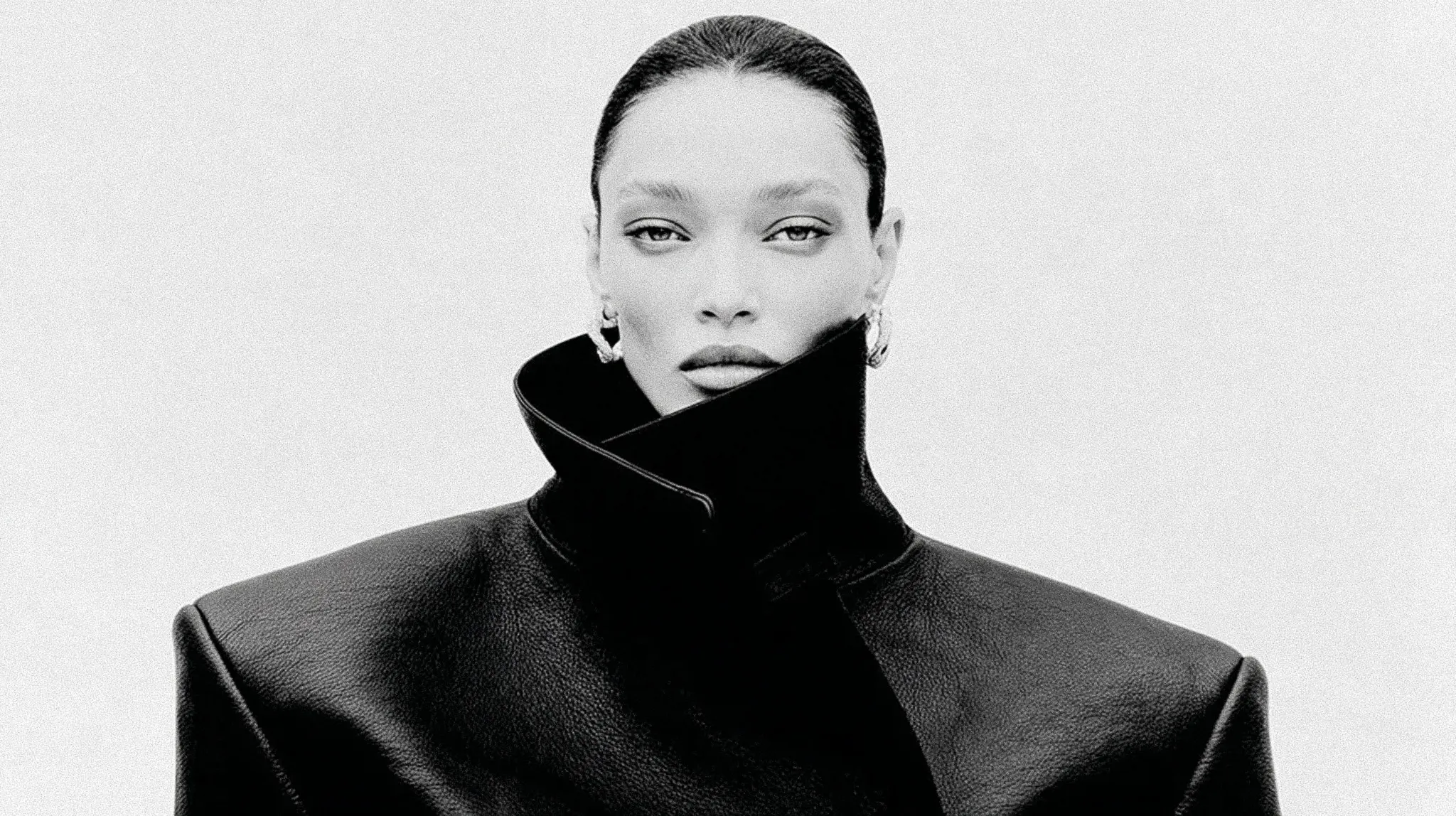 Black and white portrait of a woman with slicked-back hair wearing a high-collar leather jacket and hoop earrings.