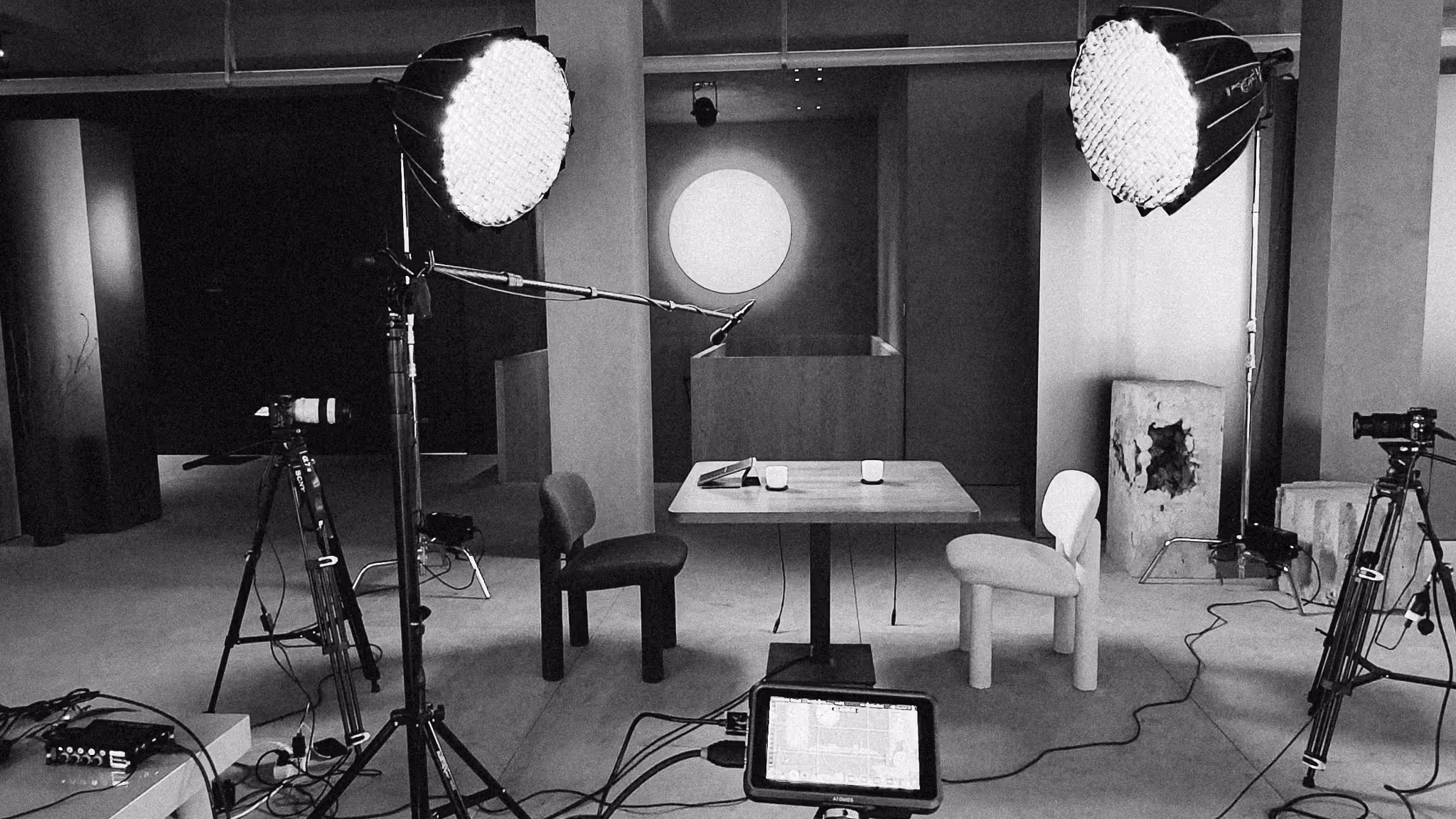 Black and white photo of a video recording setup with two chairs and a table lit by two studio lights and surrounded by cameras and cables.
