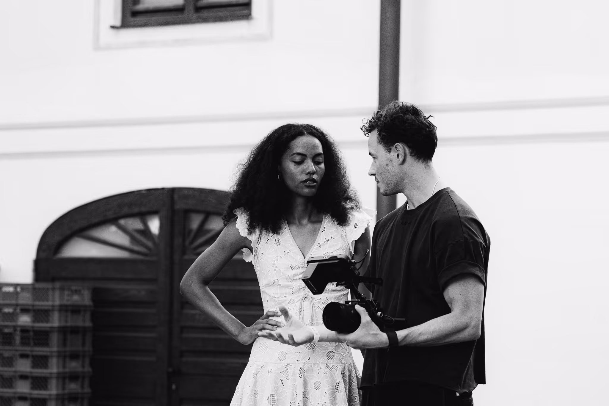 Man holding a video camera talking to a woman in a white lace dress outdoors.