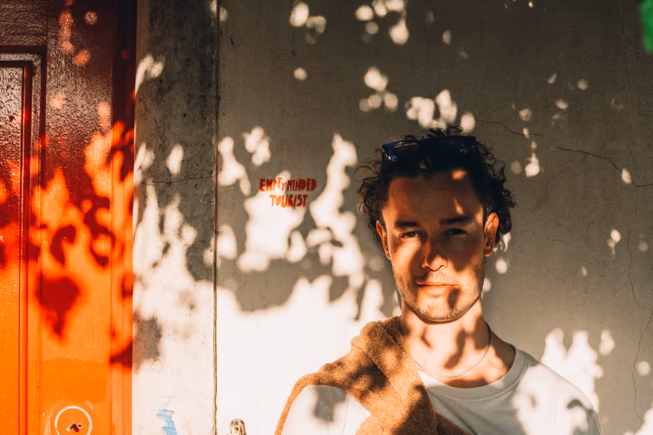 Man standing in sunlight with shadows of leaves on his face and a bright red door to the left.