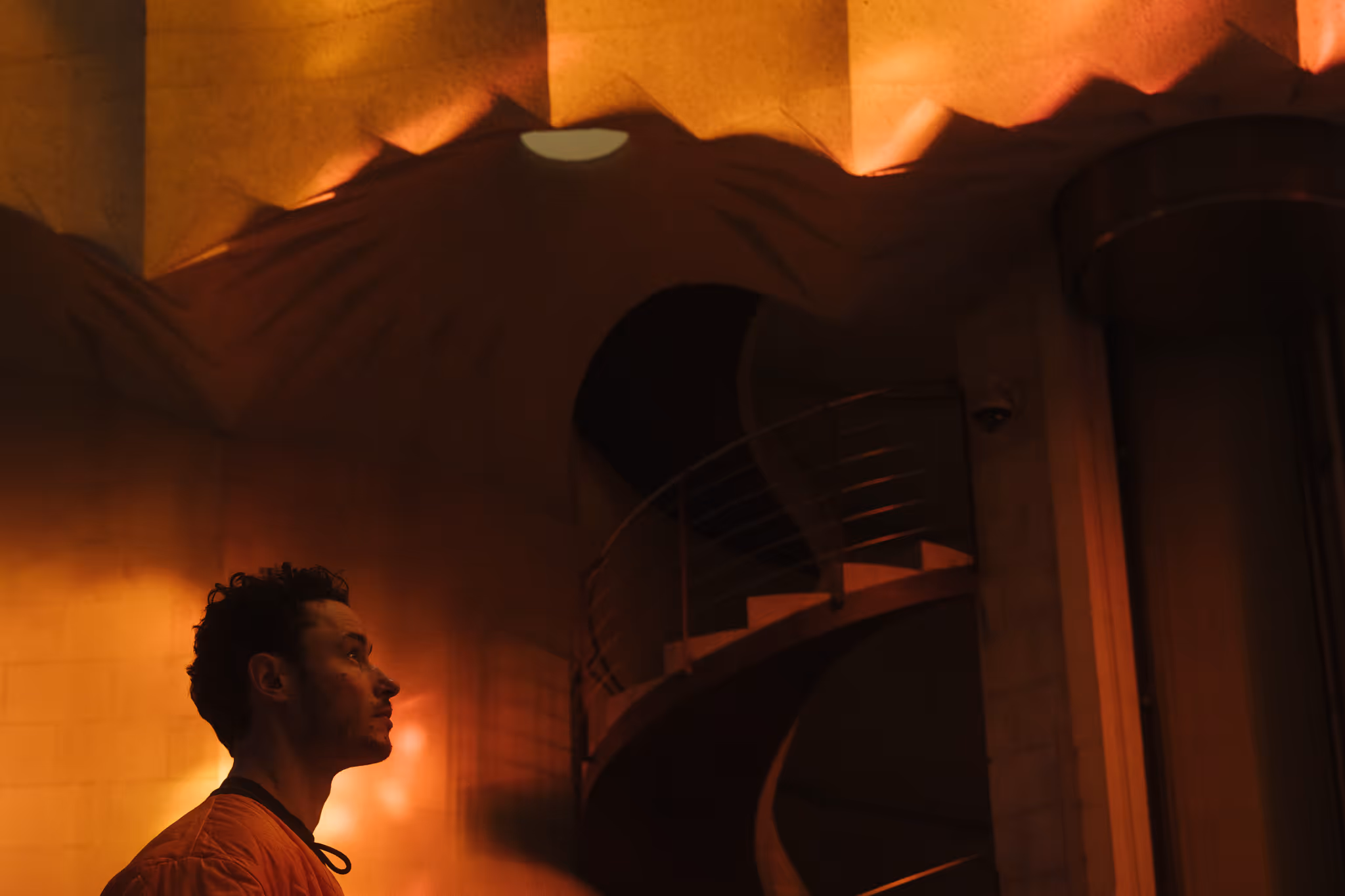 Profile of a man in a warm-toned room looking thoughtfully upward near a curved staircase.