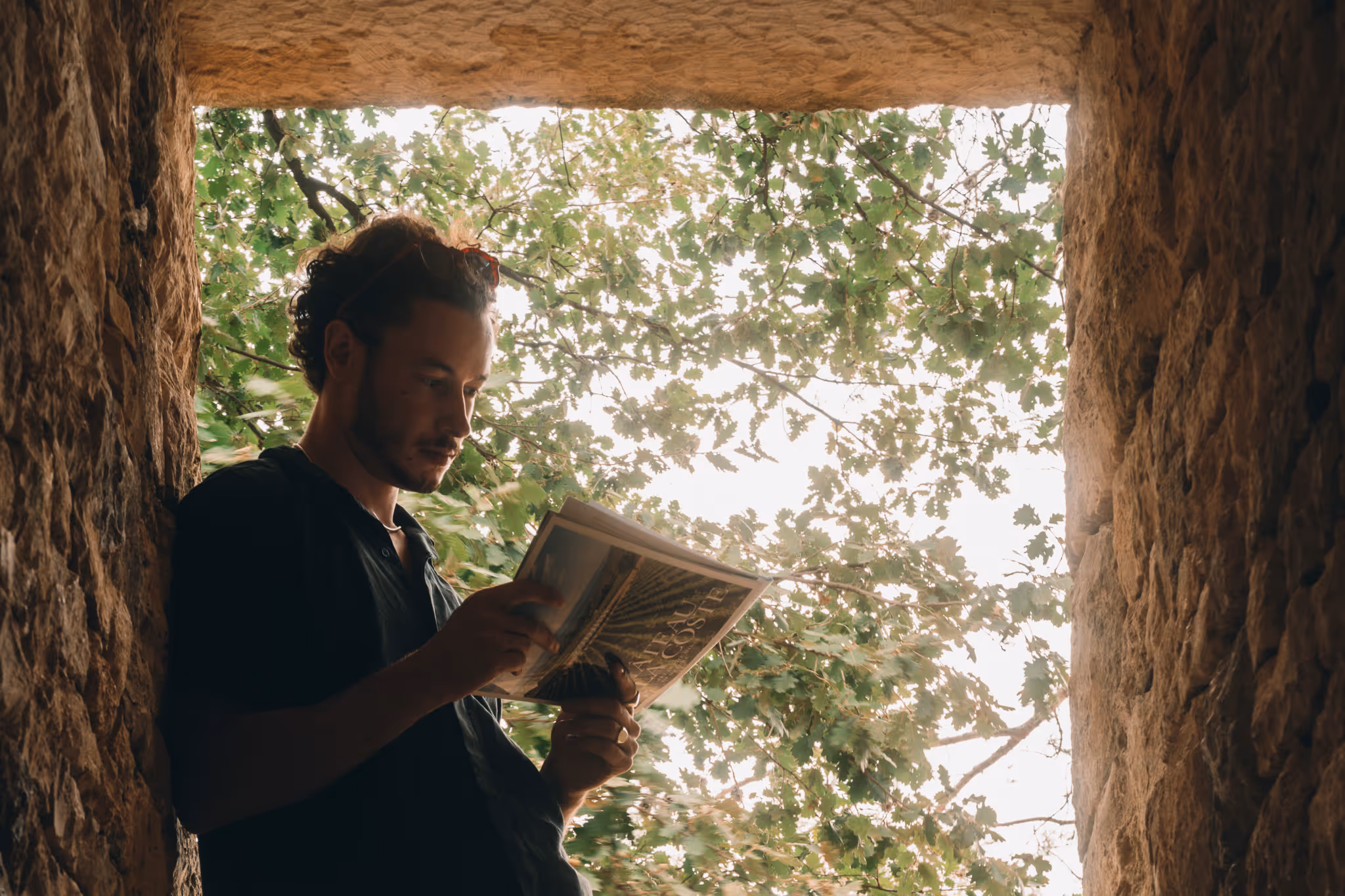Young man reading a book near a stone wall with green tree leaves visible in the background.