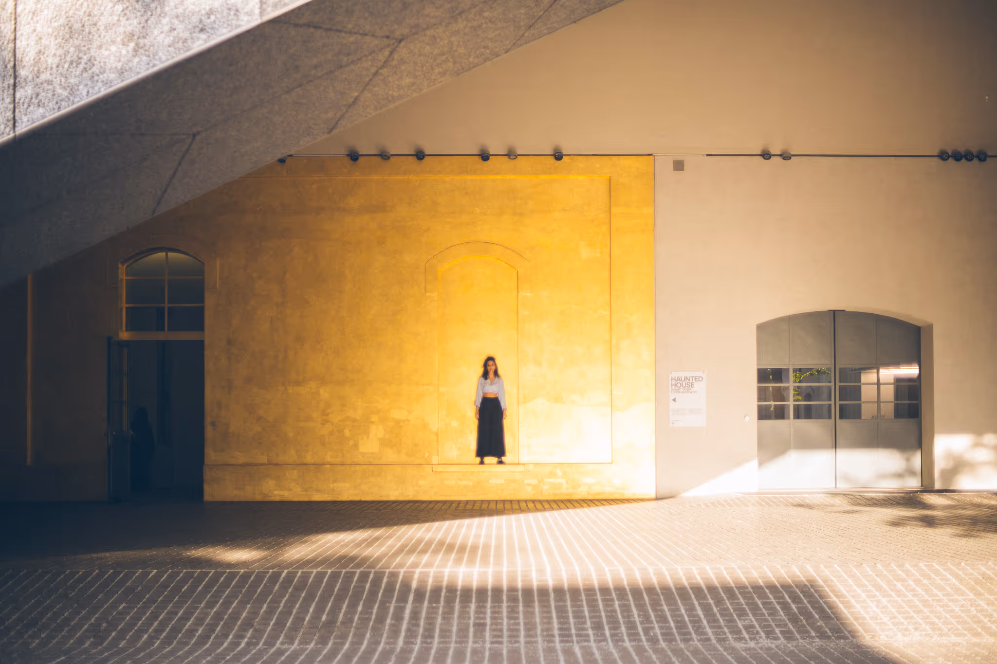 Woman in light blouse and long dark skirt standing in a recessed section of a large yellow and beige wall under natural light and shadows.