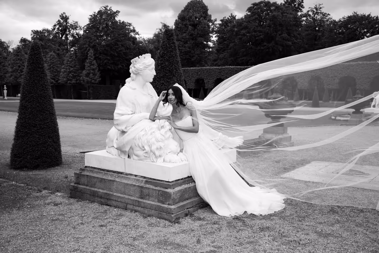 Bride in a wedding dress with a long flowing veil leaning against a large white statue of a seated woman and lion in a garden.