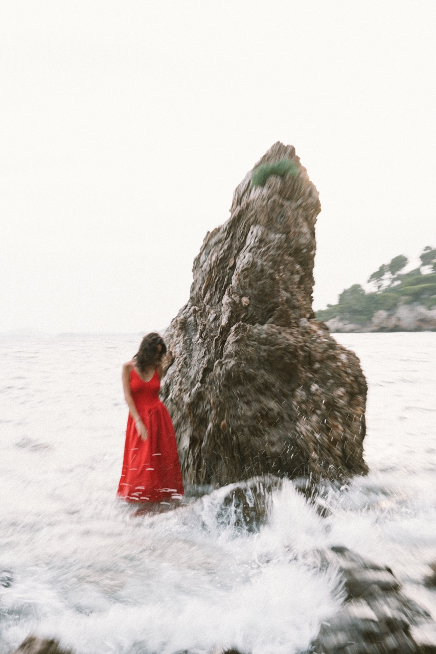 AIDA by FADETOBLACK, editorial portrait photography, image 21: A woman with dark curly hair in a red sleeveless dress poses in the sea beside a tall jagged rock, waves