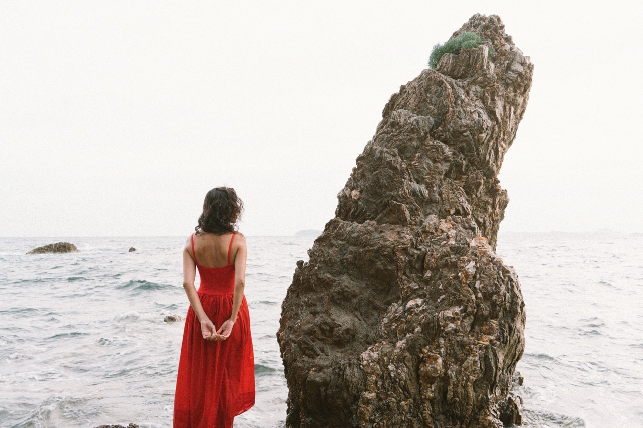 AIDA by FADETOBLACK, editorial portrait photography, image 25: A woman with dark curly hair in a red sleeveless dress poses in the sea beside a tall jagged rock, waves