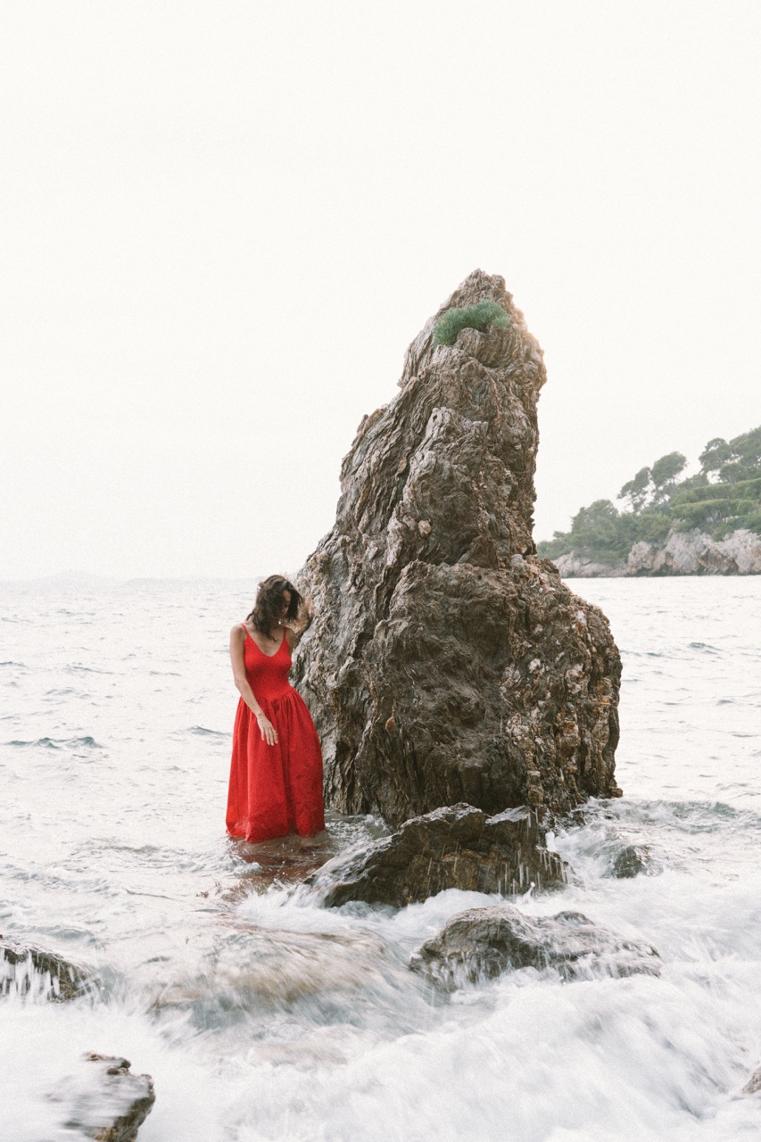 AIDA by FADETOBLACK, editorial portrait photography, image 20: A woman with dark curly hair in a red sleeveless dress poses in the sea beside a tall jagged rock, waves
