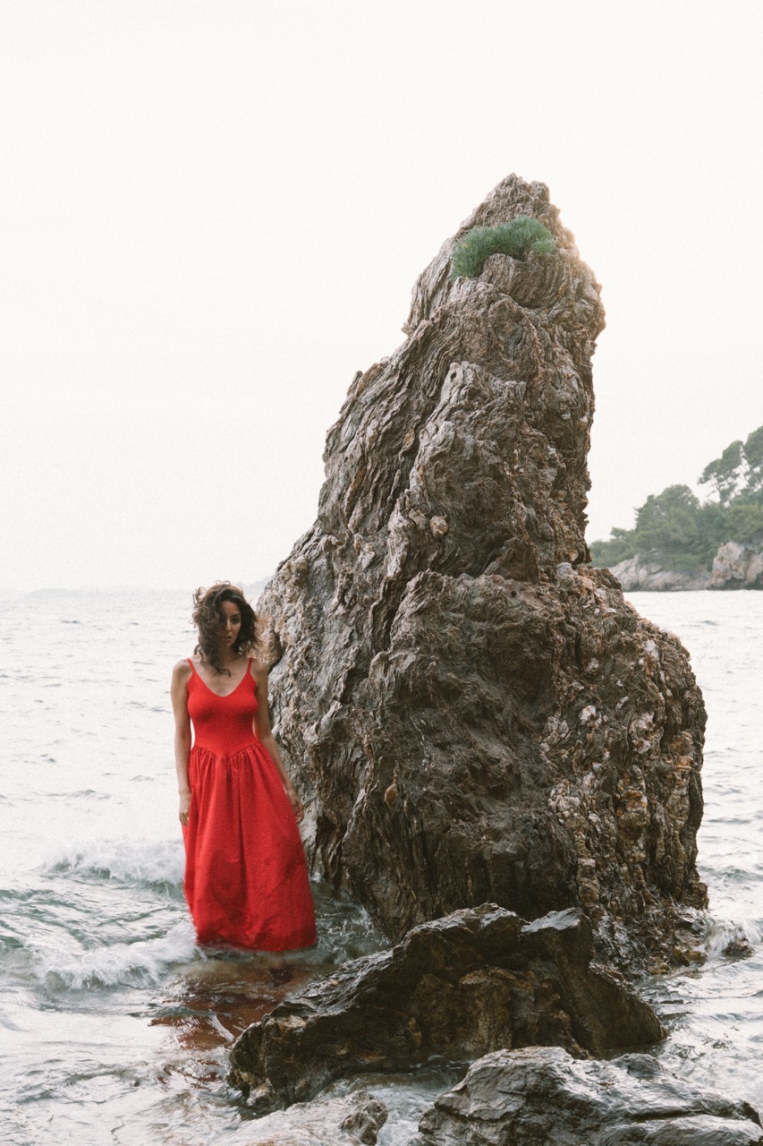 AIDA by FADETOBLACK, editorial portrait photography, image 15: A woman with dark curly hair in a red sleeveless dress poses in the sea beside a tall jagged rock, waves