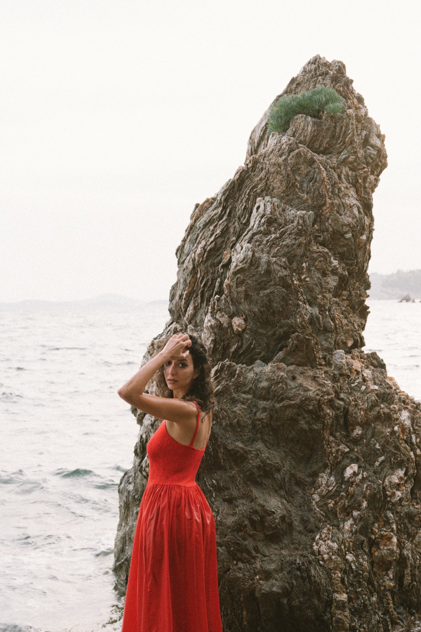 Woman in a red dress seen from behind, facing the sea beside a tall jagged rock under a pale sky. Editorial portrait photography by FADETOBLACK