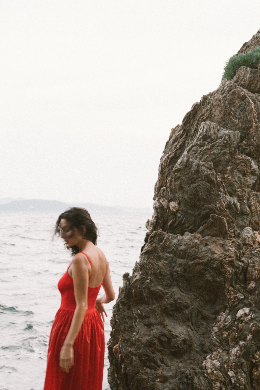 AIDA by FADETOBLACK, editorial portrait photography, image 8: A woman with dark curly hair in a red sleeveless dress poses in the sea beside a tall jagged rock, waves