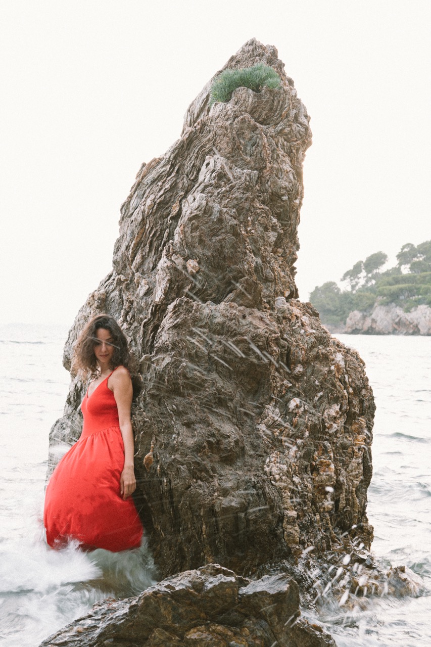 AIDA by FADETOBLACK, editorial portrait photography, image 14: A woman with dark curly hair in a red sleeveless dress poses in the sea beside a tall jagged rock, waves