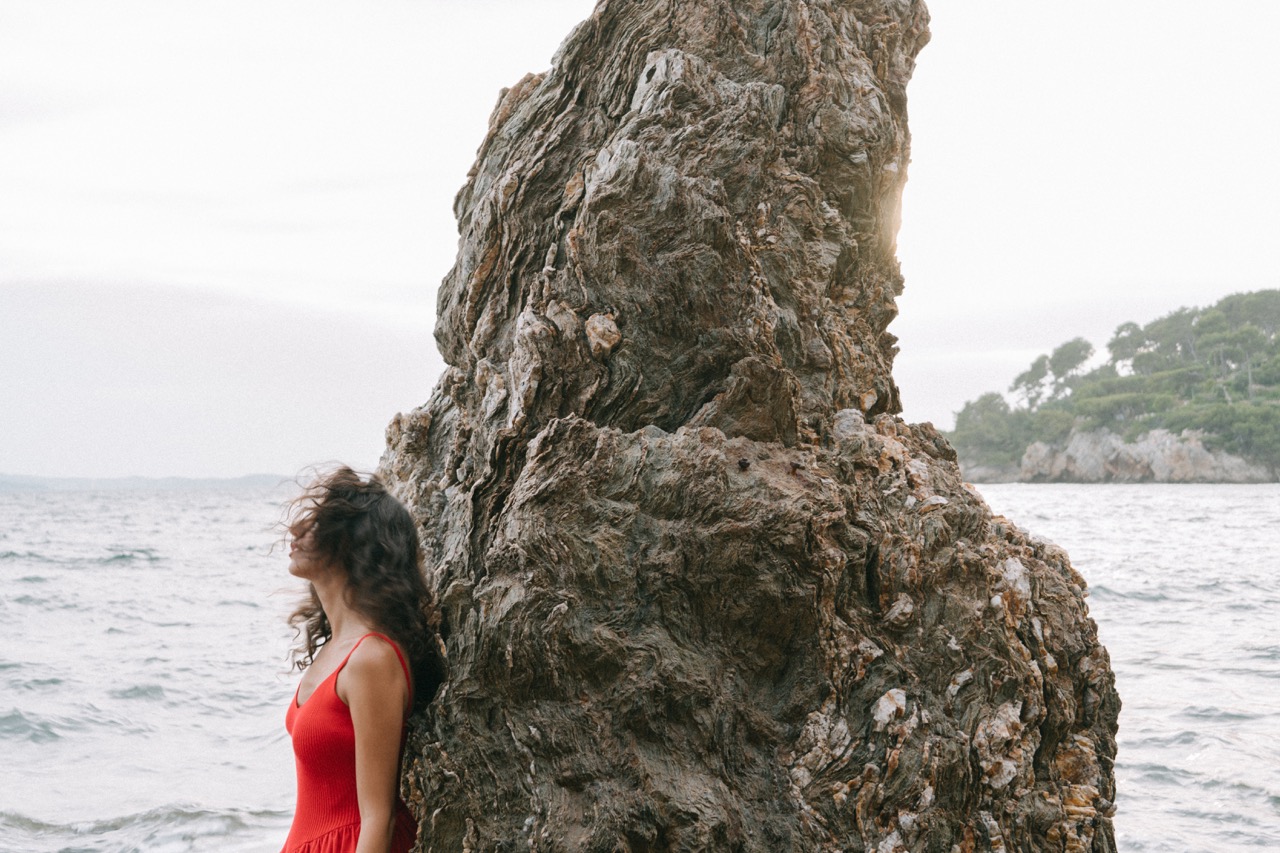AIDA by FADETOBLACK, editorial portrait photography, image 22: A woman with dark curly hair in a red sleeveless dress poses in the sea beside a tall jagged rock, waves