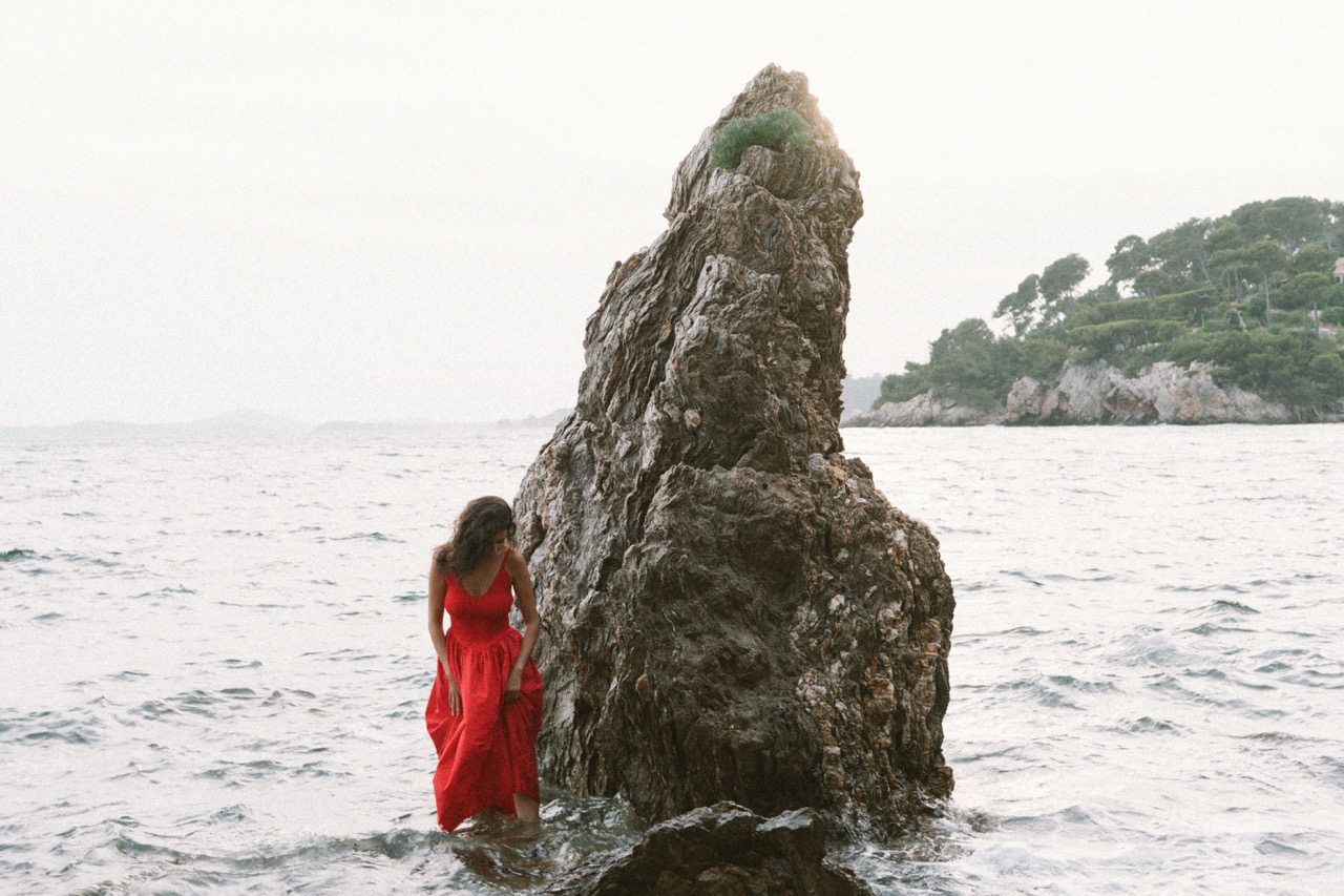 AIDA by FADETOBLACK, editorial portrait photography, image 24: A woman with dark curly hair in a red sleeveless dress poses in the sea beside a tall jagged rock, waves