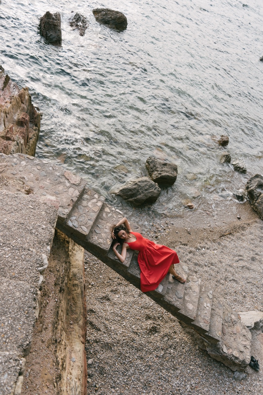 AIDA by FADETOBLACK, editorial portrait photography, image 11: A woman with dark curly hair in a red sleeveless dress poses in the sea beside a tall jagged rock, waves