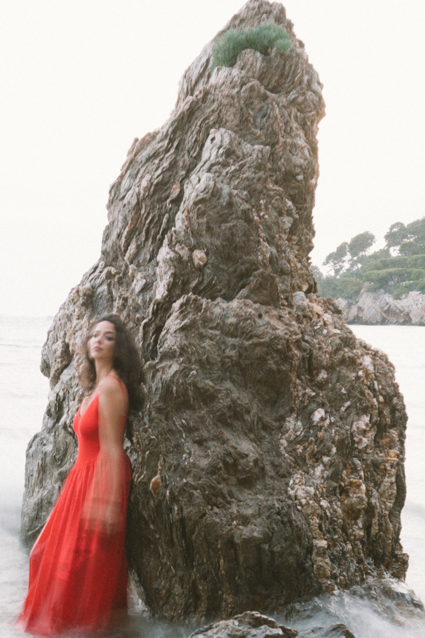 AIDA by FADETOBLACK, editorial portrait photography, image 6: A woman with dark curly hair in a red sleeveless dress poses in the sea beside a tall jagged rock, waves