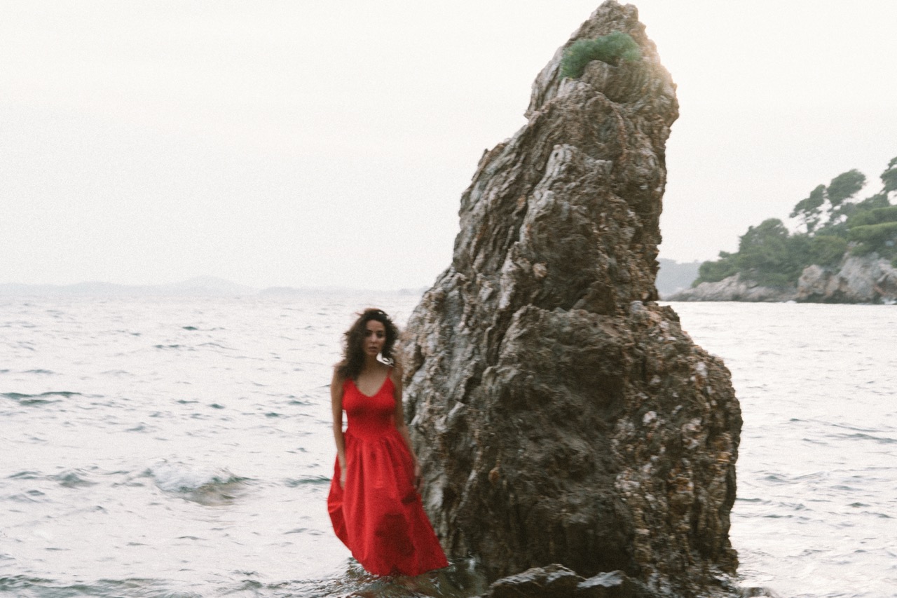 AIDA by FADETOBLACK, editorial portrait photography, image 28: A woman with dark curly hair in a red sleeveless dress poses in the sea beside a tall jagged rock, waves