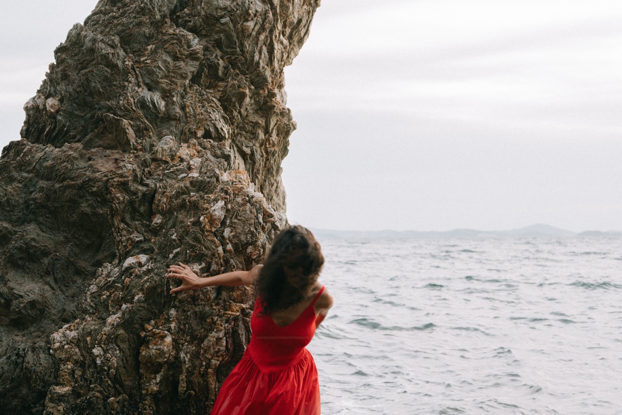 AIDA by FADETOBLACK, editorial portrait photography, image 23: A woman with dark curly hair in a red sleeveless dress poses in the sea beside a tall jagged rock, waves