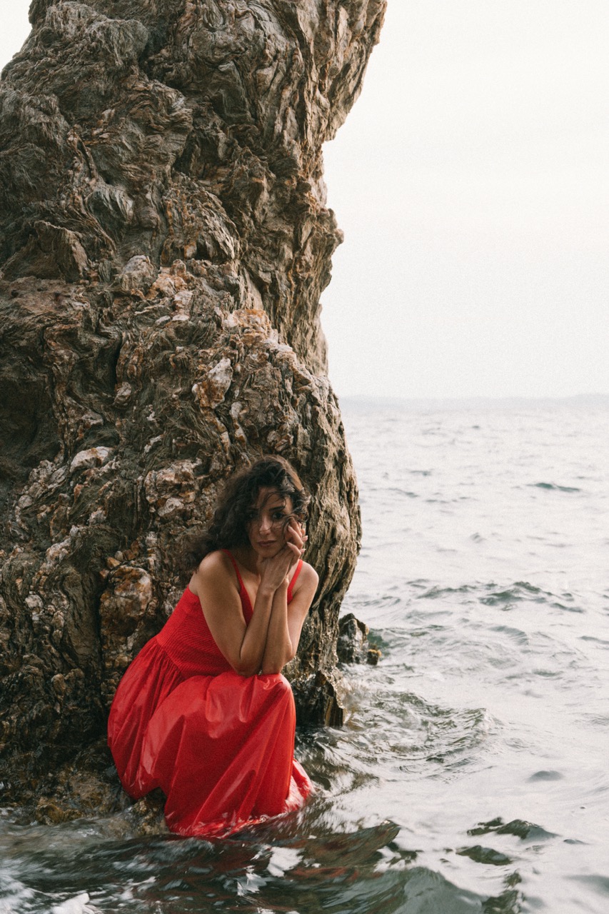 AIDA by FADETOBLACK, editorial portrait photography, image 13: A woman with dark curly hair in a red sleeveless dress poses in the sea beside a tall jagged rock, waves