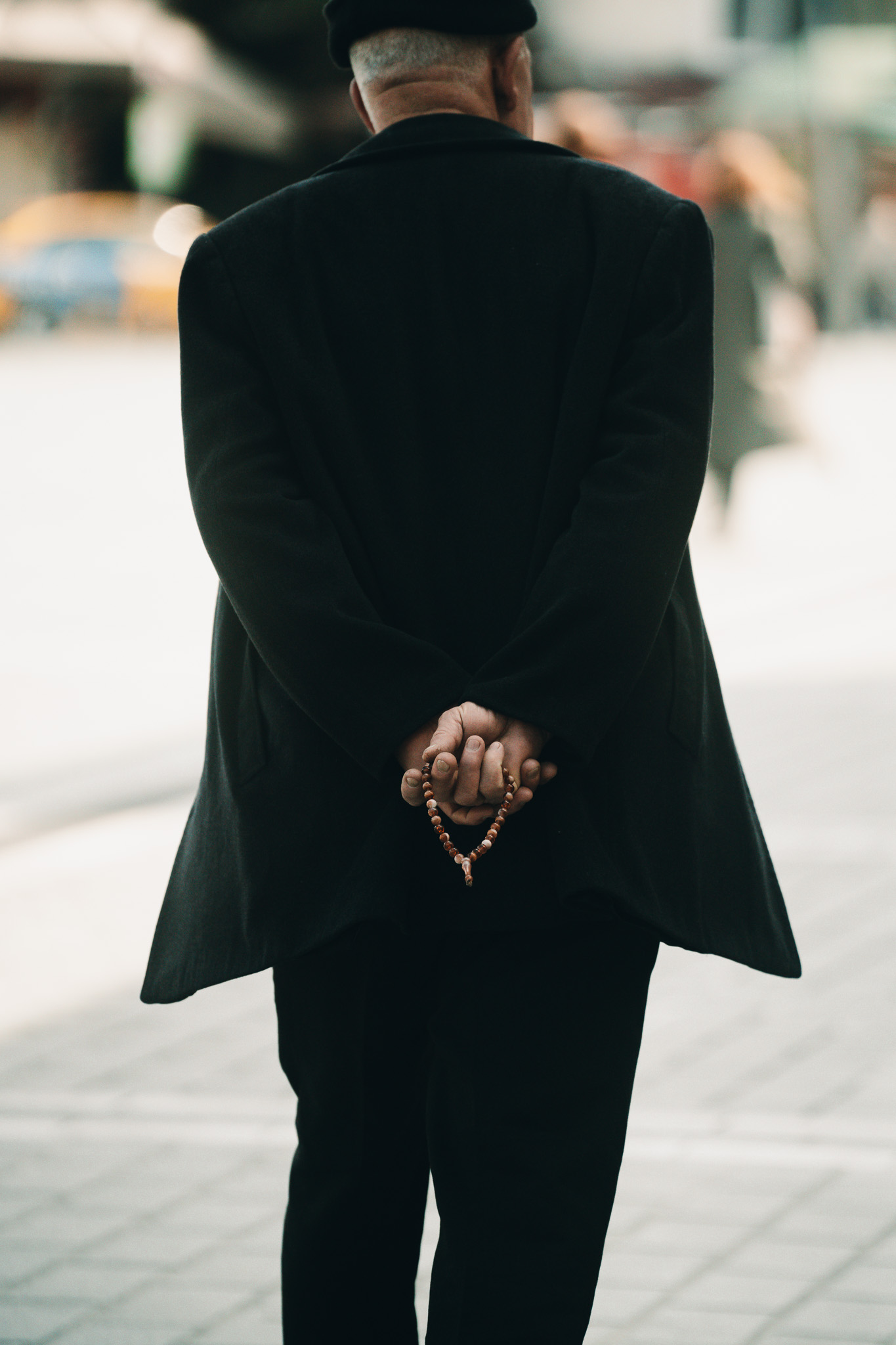 Elderly man in a black coat seen from behind, holding wooden prayer beads behind his back. Street photography by FADETOBLACK