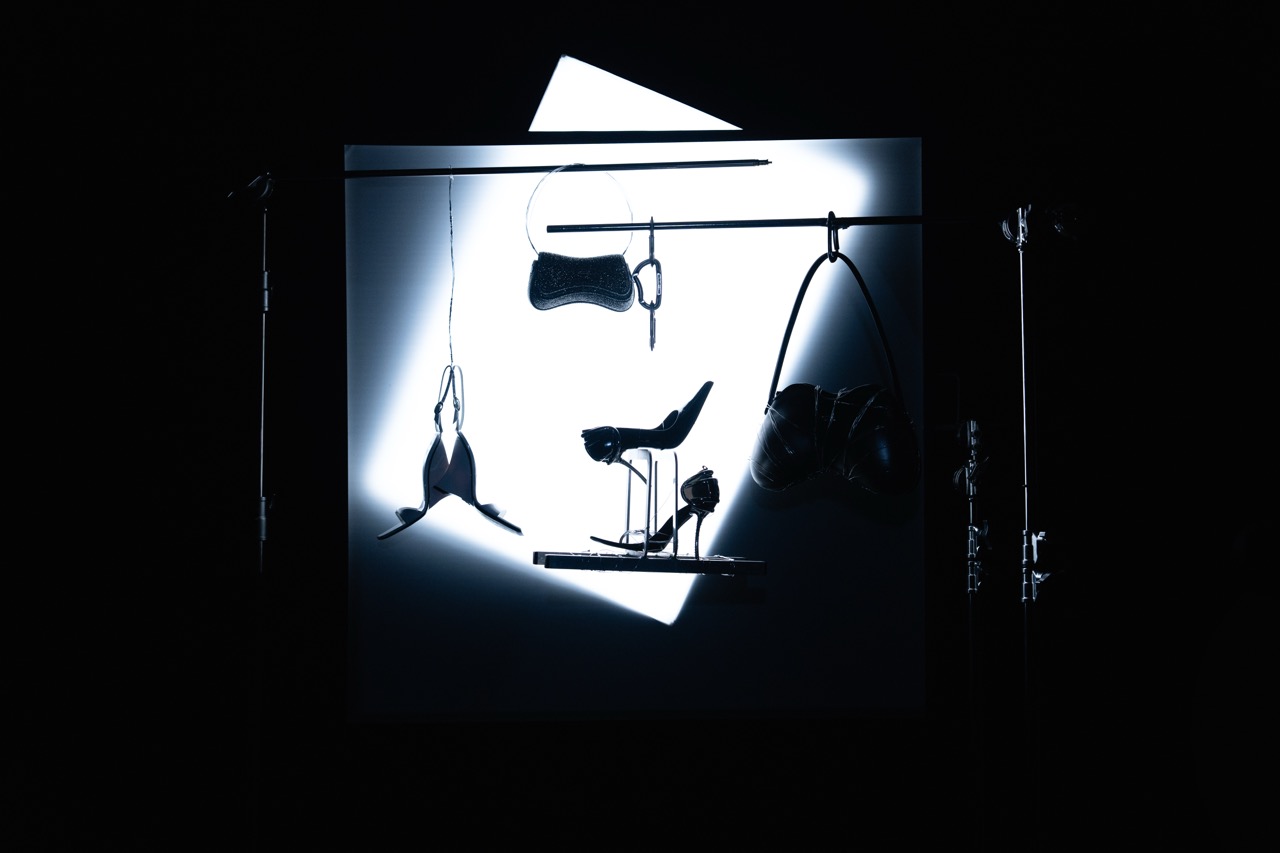 Silhouetted heels and bags hang from a steel bar in front of a glowing white lightbox in darkness. Still life campaign photography by FADETOBLACK